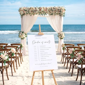 May include: A beach wedding ceremony setup with a white sign displaying the order of events. The sign is on a wooden easel, with wooden chairs and floral arrangements. A floral arch and sheer fabric frame the ocean backdrop.
