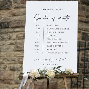 May include: A white wedding sign on a wooden easel, displaying the order of events for Amanda and Steven's wedding. The sign includes the date, time, and activities, with floral and ribbon accents at the base.