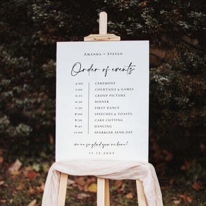 May include: A white wedding sign on a wooden easel, displaying the text "Amanda + Steven" and "Order of events." The schedule includes ceremony, dinner, dancing, and sparkler send-off. The date is 11.17.2025.