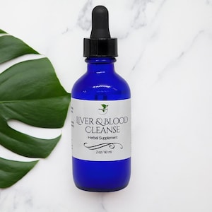 Può includere: Una bottiglia di vetro blu cobalto con un contagocce nero. L'etichetta recita "LIVER & BLOOD CLEANSE Herbal Supplement" con un logo verde. La bottiglia contiene 60 ml di liquido. Una grande foglia verde è sullo sfondo.