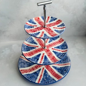 Puede incluir: Un soporte de tres niveles con platos de cerámica redondos decorados con un patrón de Union Jack rojo y azul hecho de pequeños puntos. Los platos están conectados por una varilla central de metal plateado con un asa en forma de T.
