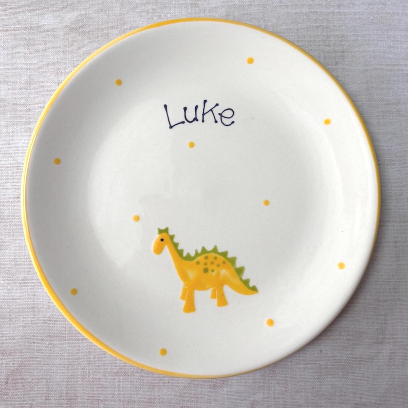 May include: A round white ceramic plate with a yellow rim, featuring a yellow dinosaur illustration with green spikes and the name Luke written in dark blue script. The plate is decorated with small yellow dots scattered across the surface.