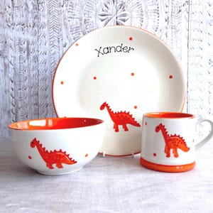 May include: A ceramic dinnerware set consisting of a plate, a bowl, and a mug. Each piece is white with a bright orange interior and rim, featuring a hand-painted orange dinosaur motif and orange polka dots. The plate is personalized with the name Xander.