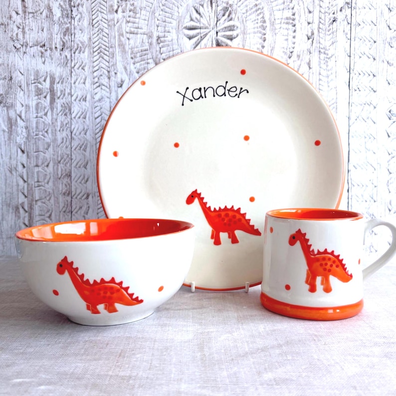 May include: A ceramic dinnerware set consisting of a plate, a bowl, and a mug. Each piece is white with a bright orange interior and rim, featuring a hand-painted orange dinosaur motif and orange polka dots. The plate is personalized with the name Xander.