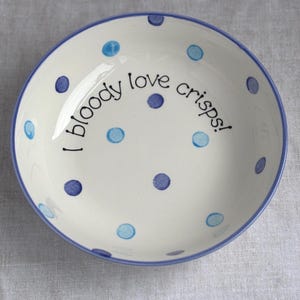 May include: A round ceramic bowl with a cream-coloured base, featuring a blue rim and a pattern of scattered blue polka dots. The interior of the bowl displays the black handwritten text "I bloody love crisps!" centred across the middle.