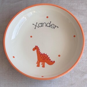 May include: A round ceramic bowl with an off-white interior and a bright orange rim. The center features a hand-painted orange dinosaur graphic, small orange polka dots, and the name Xander written in black cursive script across the top.