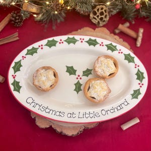 May include: An oval white ceramic serving platter with a red trim and green holly leaf and berry design. The platter holds three mince pies and the text "Christmas at Little Orchard".