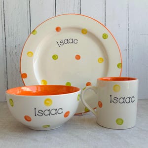 Può includere: Un set di stoviglie in ceramica composto da un piatto, una ciotola e una tazza, tutti con base color crema e pois arancioni e gialli. Ogni pezzo è personalizzato con il nome Isaac in caratteri neri. La ciotola e la tazza hanno interni arancioni.