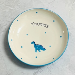 May include: A round ceramic plate with an off-white base, featuring a blue dinosaur illustration in the center and the name Thomas written in black script. The plate is decorated with small blue polka dots and has a blue rim around the edge.