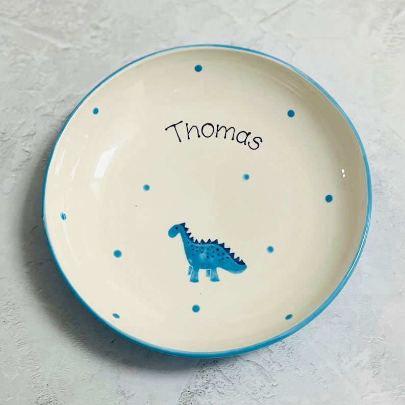 May include: A round ceramic plate with an off-white base, featuring a blue dinosaur illustration in the center and the name Thomas written in black script. The plate is decorated with small blue polka dots and has a blue rim around the edge.