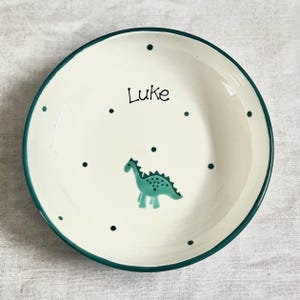 May include: A round ceramic bowl with an off-white base and a dark green rim. The interior features a pattern of small dark green dots, a hand-painted teal dinosaur illustration, and the name Luke written in black script lettering.