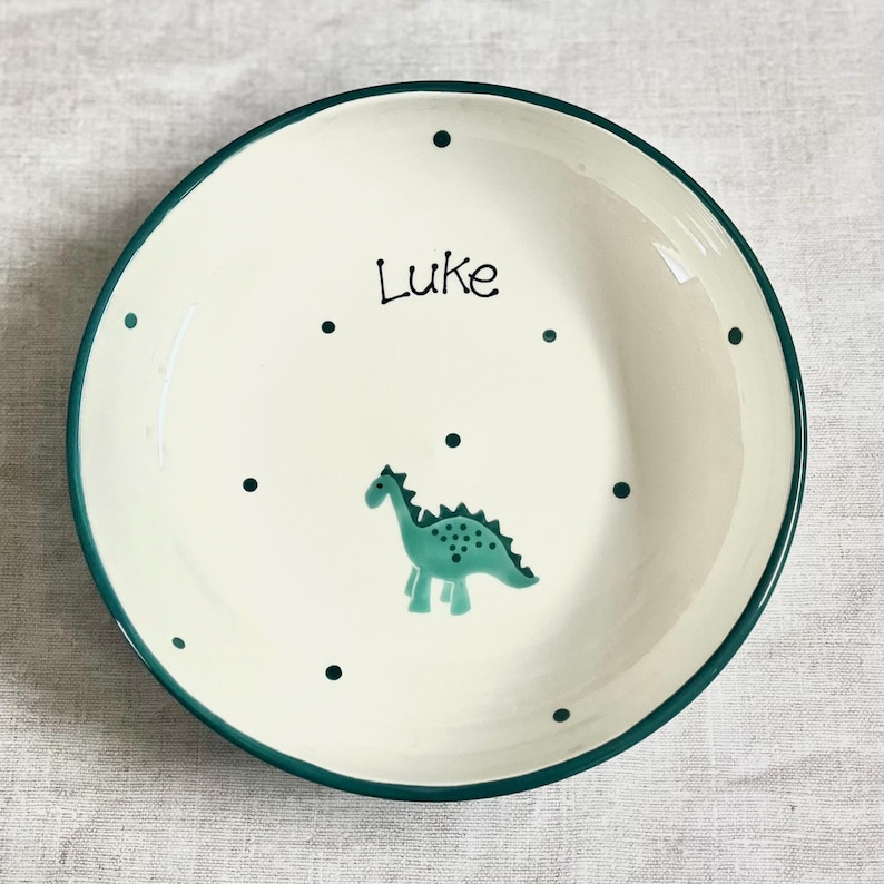 May include: A round ceramic bowl with an off-white base and a dark green rim. The interior features a pattern of small dark green dots, a hand-painted teal dinosaur illustration, and the name Luke written in black script lettering.