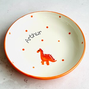 May include: A round ceramic bowl with a white interior and an orange rim. The bowl features a hand-painted orange dinosaur graphic and scattered orange polka dots. The name Arthur is written in black script lettering on the interior surface.