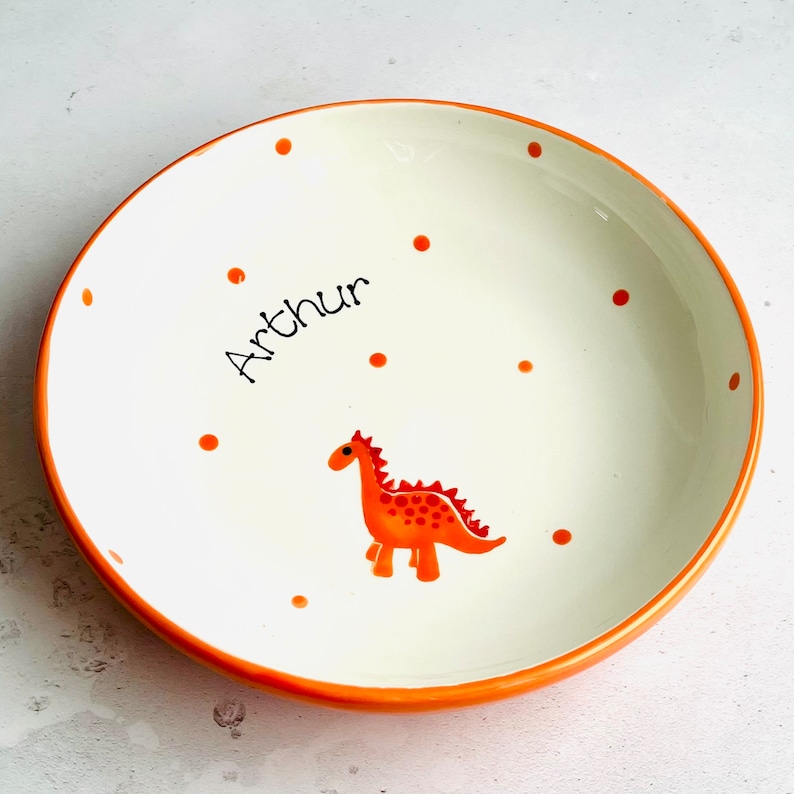 May include: A round ceramic bowl with a white interior and an orange rim. The bowl features a hand-painted orange dinosaur graphic and scattered orange polka dots. The name Arthur is written in black script lettering on the interior surface.