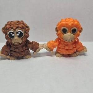 May include: Two small, detailed monkey figurines. One is brown, the other is orange. Both have large, expressive eyes and are holding hands. The figurines are made of a textured material and have a cartoonish appearance.