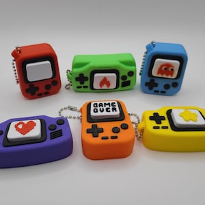 May include: A collection of six colorful, retro-style handheld game console keychains. Each keychain features a different color, including red, green, blue, purple, orange, and yellow, with a unique icon on the screen. The orange keychain has the text "GAME OVER".