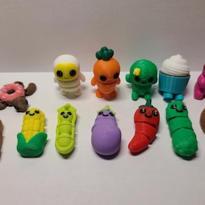 May include: A collection of colorful, miniature food and character figurines. Includes a donut, hot dog, corn, caterpillar, eggplant, pepper, and a cupcake. Other figures include a bear, an astronaut, and a carrot.