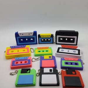 May include: A collection of colorful retro-style keychains shaped like cassette tapes and floppy disks. The keychains feature a variety of bright colors, including blue, yellow, pink, green, and black. Each keychain has a small metal chain attached.