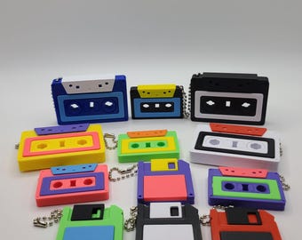 May include: A collection of colorful retro-style keychains shaped like cassette tapes and floppy disks. The keychains feature a variety of bright colors, including blue, yellow, pink, green, and black. Each keychain has a small metal chain attached.
