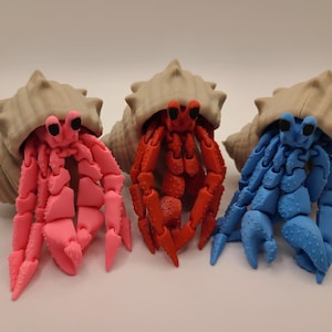 May include: Three colorful hermit crab figurines. A pink crab, a red crab, and a blue crab are each emerging from a beige seashell. The crabs have articulated legs and claws, and black eyes. The seashells have a textured surface.