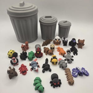 May include: Three gray plastic trash cans of varying sizes arranged behind a collection of small, colorful animal-shaped erasers. The erasers include various creatures like a ladybug, squirrel, scorpion, spider, butterfly, and other wildlife figures.