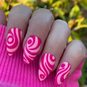 May include: A hand with long, almond-shaped nails. Each nail is painted with a bright pink base and a swirling pattern in a darker shade of pink. The nails are set against a bright pink background.