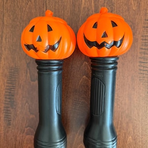 May include: Two identical handheld flashlights featuring bright orange plastic pumpkin toppers with black painted jack-o'-lantern faces. Each pumpkin is attached to a cylindrical black plastic handle with a ribbed grip section and a flared base.