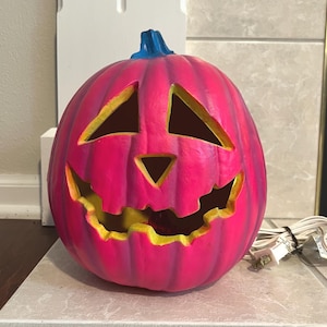May include: A vibrant pink ceramic jack-o'-lantern with a carved face and a blue stem. The interior of the face is yellow. The electric cord is white.