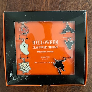 May include: A set of six Halloween glassware charms in a box. The charms are made of metal and feature a variety of Halloween designs, including a pumpkin, a ghost, a spider web, a bat, a black cat, and a witch. The box is orange and black and has the words "Halloween Glassware Charms" printed on it.