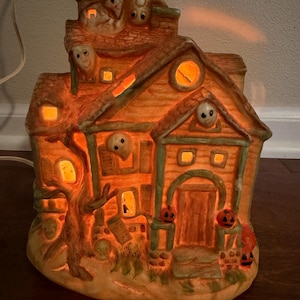 May include: A ceramic Halloween haunted house figurine with a warm orange internal glow. The structure features multiple ghosts peering from windows and the roof, small jack-o'-lanterns flanking the front door, and a gnarled tree on the left side.
