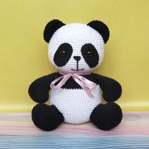 May include: A crocheted panda bear plush toy with black ears, eyes, and paws. The panda is wearing a pink ribbon around its neck and is sitting on a colorful wooden surface.