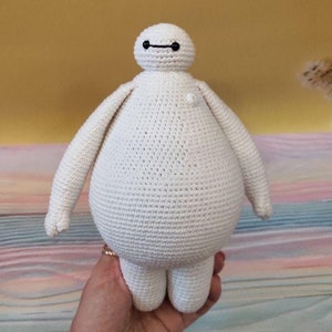 May include: White crocheted plush toy resembling Baymax from the Disney movie Big Hero 6. The toy has a round body, short legs, and a simple face with black eyes and a black mouth.
