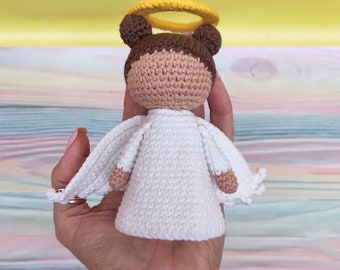 May include: A crocheted angel doll with a white dress, brown hair, and a yellow halo. The doll is being held in a person's hand.