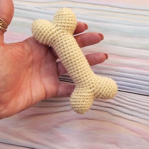 May include: A crocheted dog bone toy in a cream color. The bone is held in a hand against a light pink and blue striped background.