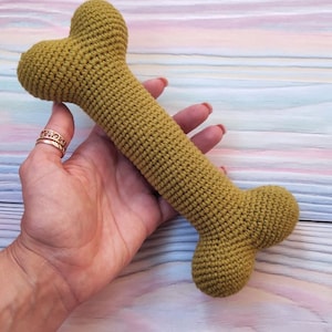 Op de afbeelding: Een gehaakt hondenbot speelgoed in een gedempte groene kleur. Het bot wordt in een hand gehouden tegen een gestreepte achtergrond.