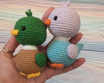 May include: Two crocheted stuffed animals, a green duck and a light blue and purple bird. The duck has a brown body and orange feet. The bird has orange feet and a light purple head.
