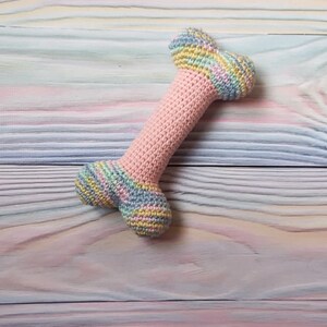 Op de afbeelding: Een gehaakt hondenbot speelgoed in roze, blauw en geel op een pastelkleurige houten achtergrond.