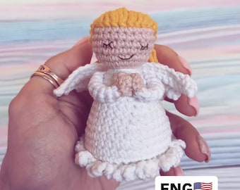 Könnte beinhalten: Eine gehäkelte Engelpuppe mit einem weißen Kleid, gelben Haaren und geschlossenen Augen. Die Puppe wird in einer Hand gehalten. Der Text "Pattern" und "ENG" ist auf dem Bild zu sehen.