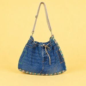 Pode incluir: Uma bolsa de ombro em ganga azul feita a partir de calças de ganga reutilizadas, com fecho de cordão entrançado, alça bege ajustável e acabamentos decorativos tecidos em tons de verde e castanho nas laterais e base.