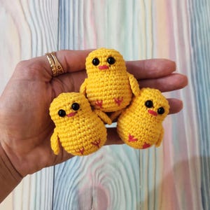 May include: Three small crocheted yellow chicks with black eyes and red beaks. The chicks are sitting in a person's hand.