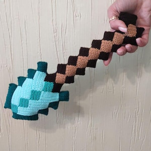 May include: Crocheted toy sword in a pixelated design. The blade is light blue and teal, while the handle alternates brown and tan squares. The toy is held against a textured, off-white wall.