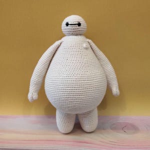 May include: White crocheted plush toy resembling Baymax from the Disney movie Big Hero 6. The toy has a round body, small arms, and legs, and a simple black face with two eyes.
