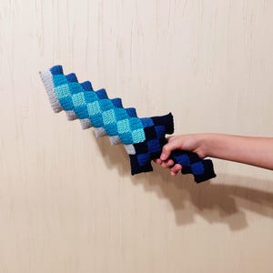 May include: A handmade crochet sword in shades of blue, gray, and navy. The sword features a pixelated design, with a checkered pattern on the blade and handle. The toy is held by a person.