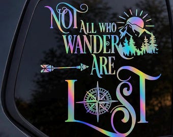 May include: An iridescent holographic vinyl decal applied to a car window featuring the text NOT ALL WHO WANDER ARE LOST. The design includes a mountain range with a sun, a horizontal arrow, and a compass rose integrated into the letter O.