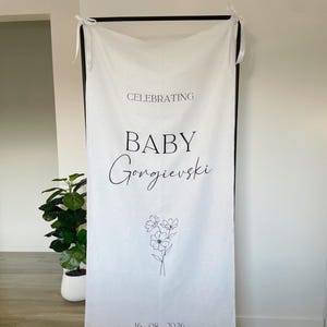 May include: A white fabric banner hanging from a black metal frame with white ribbon ties. The banner features black text that reads CELEBRATING BABY Gongievski, a black line drawing of a flower bouquet, and the date 16.08.2026 printed at the bottom.