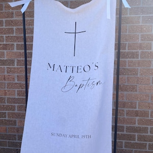 May include: A vertical off-white fabric banner suspended from a black metal frame by white ribbon ties. The banner features a black cross icon, the text MATTEO'S Baptism, and the date SUNDAY APRIL 19TH printed in black ink on the front surface.