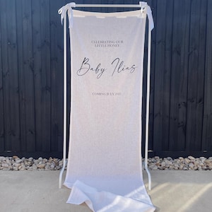 May include: A tall, off-white fabric banner suspended from a white metal frame with tied ribbon accents. The banner features black printed text that reads: CELEBRATING OUR LITTLE HONEY, Baby Ilias, and COMING JULY 2025. The fabric pools at the base.
