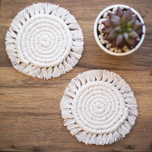 May include: Two white crocheted coasters with fringe detailing. The coasters are round and have a concentric circle design. The coasters are on a wooden surface.