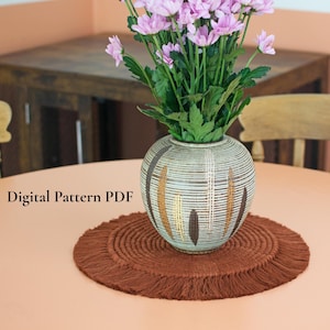 May include: A brown macrame placemat with a fringed edge sits on a peach-coloured table. A decorative vase with a striped pattern and brown and gold accents holds a bouquet of pink flowers. The text "Digital Pattern PDF" is visible.