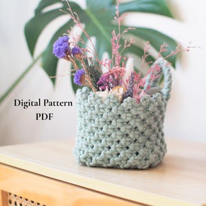 May include: A pale green, hand-crocheted basket containing dried flowers in purple, pink, and white. The basket is square with a textured stitch and a handle. Text on the left side reads "Digital Pattern PDF".